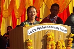 [ai] A woman speaking at a podium decorated with marigold flowers, with a sign reading 'Sankhu Palubari Community School.' In the background, a man listens attentively, while colorful drapery decorates the setting.